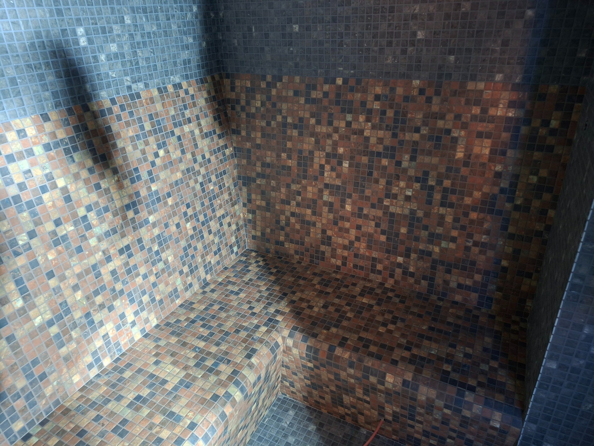 Mozaika w odcieniach brązu i szarości wyłożona w kabinie prysznicowej z siedziskiem. Widoczne fugi i załamania ścian. Górna część ściany wykończona ciemniejszą mozaiką.