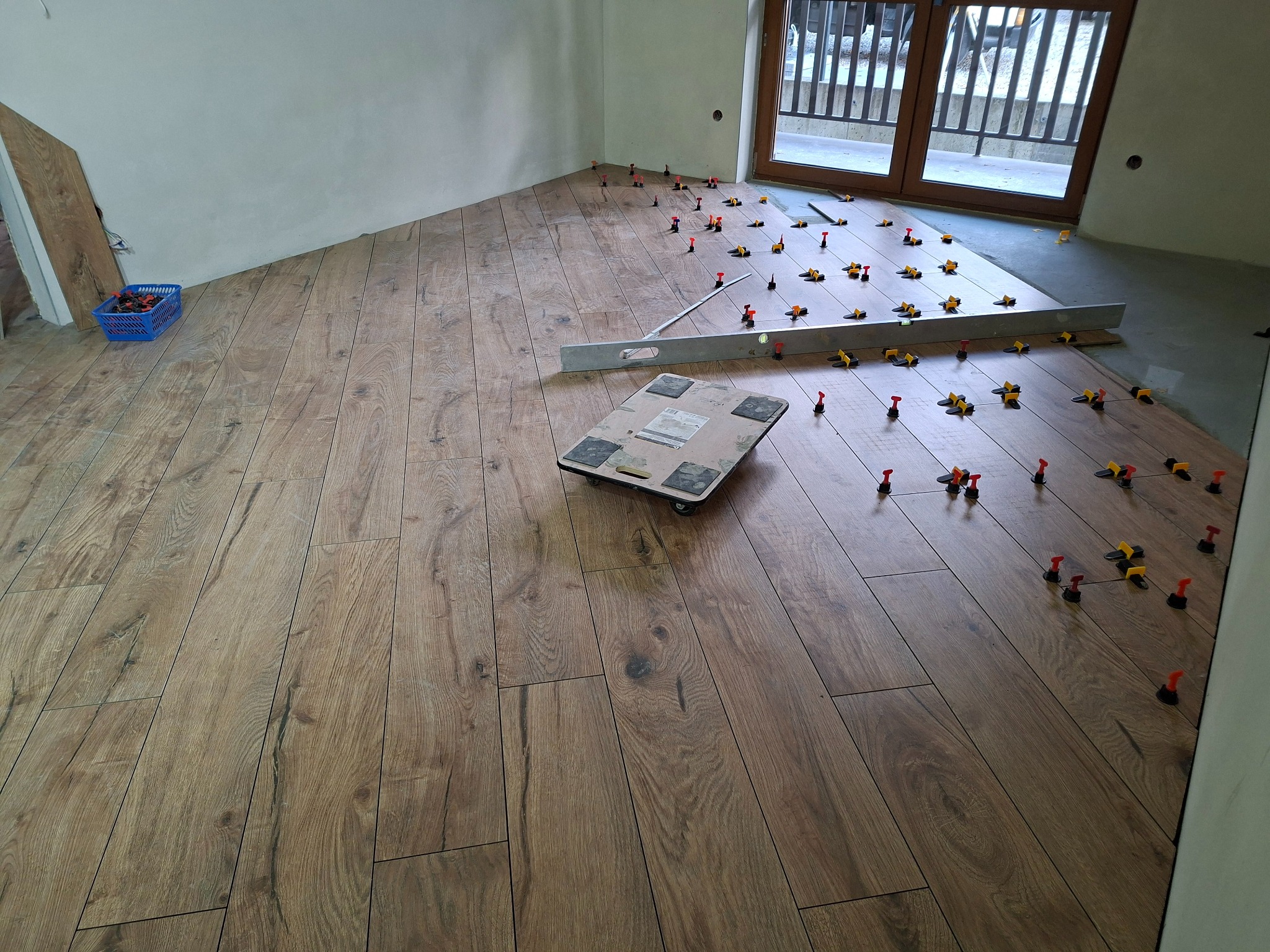 Układanie drewnopodobnych płytek podłogowych w pokoju. Widoczne poziomice, wózki transportowe i system poziomowania płytek. Częściowo ułożona podłoga, surowy stan ścian.