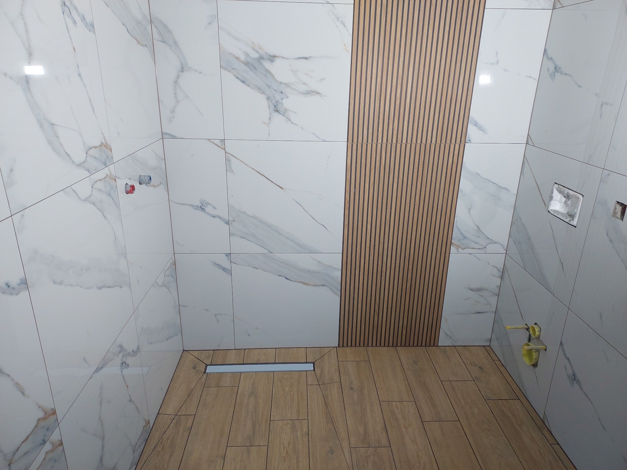 Wykończona kabina prysznicowa z białymi płytkami imitującymi marmur, drewnopodobną podłogą i panelem dekoracyjnym z pionowych listew. Widoczny odpływ liniowy.