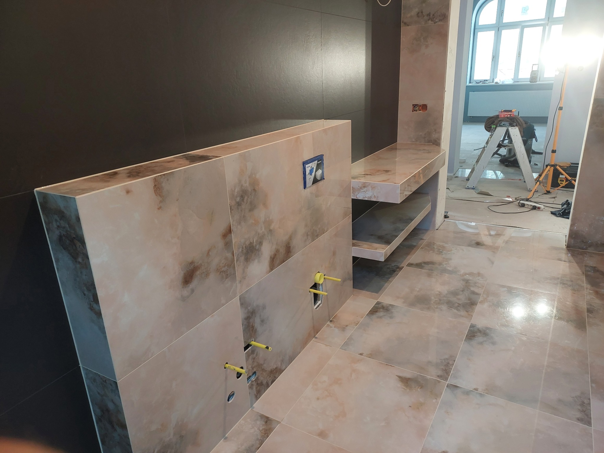 Łazienka w trakcie wykończenia: beżowe płytki imitujące marmur na podłodze i zabudowie WC, czarna ściana, widoczne rury instalacyjne i drabina w tle.