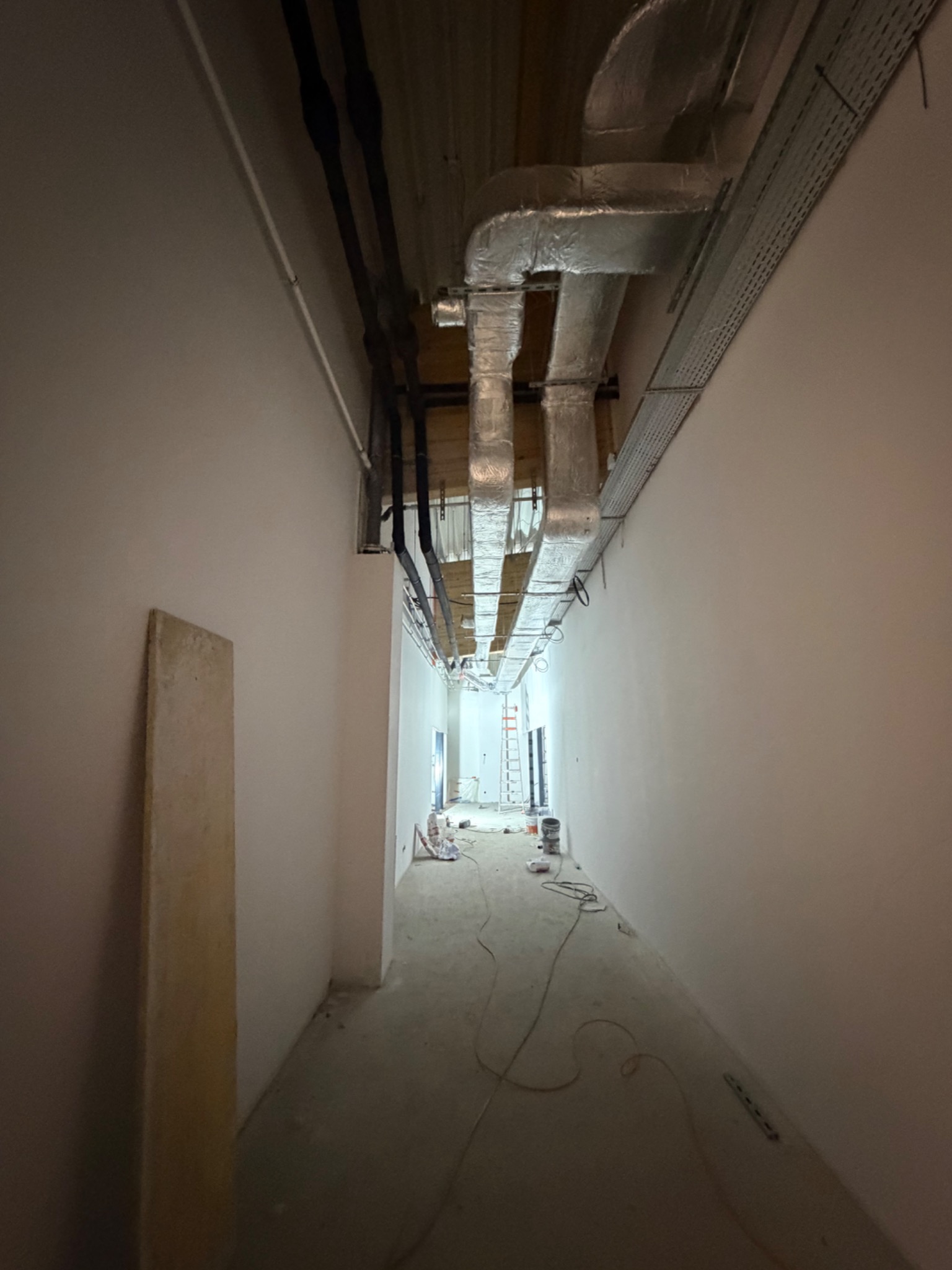 Korytarz w trakcie montażu instalacji wentylacyjnej i elektrycznej. Widoczne rury wentylacyjne pod sufitem, koryta kablowe oraz przewody na podłodze. Ściany pomalowane na biało.