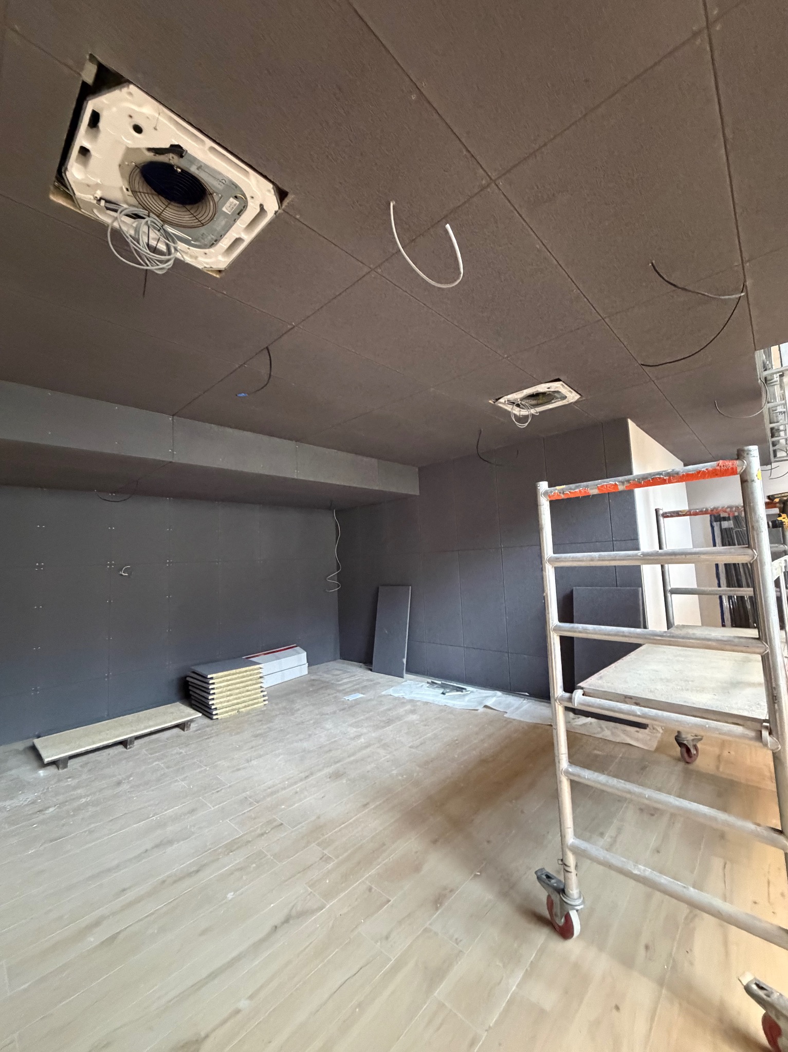 Montaż klimatyzacji kasetonowej w pomieszczeniu z szarą ścianą i podwieszanym sufitem. Widoczne rusztowanie i materiały izolacyjne.