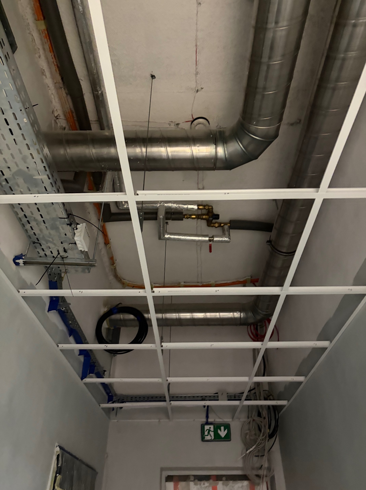 Widok na sufit podwieszany z widocznymi rurami wentylacyjnymi, kablami i elementami instalacji. Metalowe rury i kable prowadzone pod sufitem.