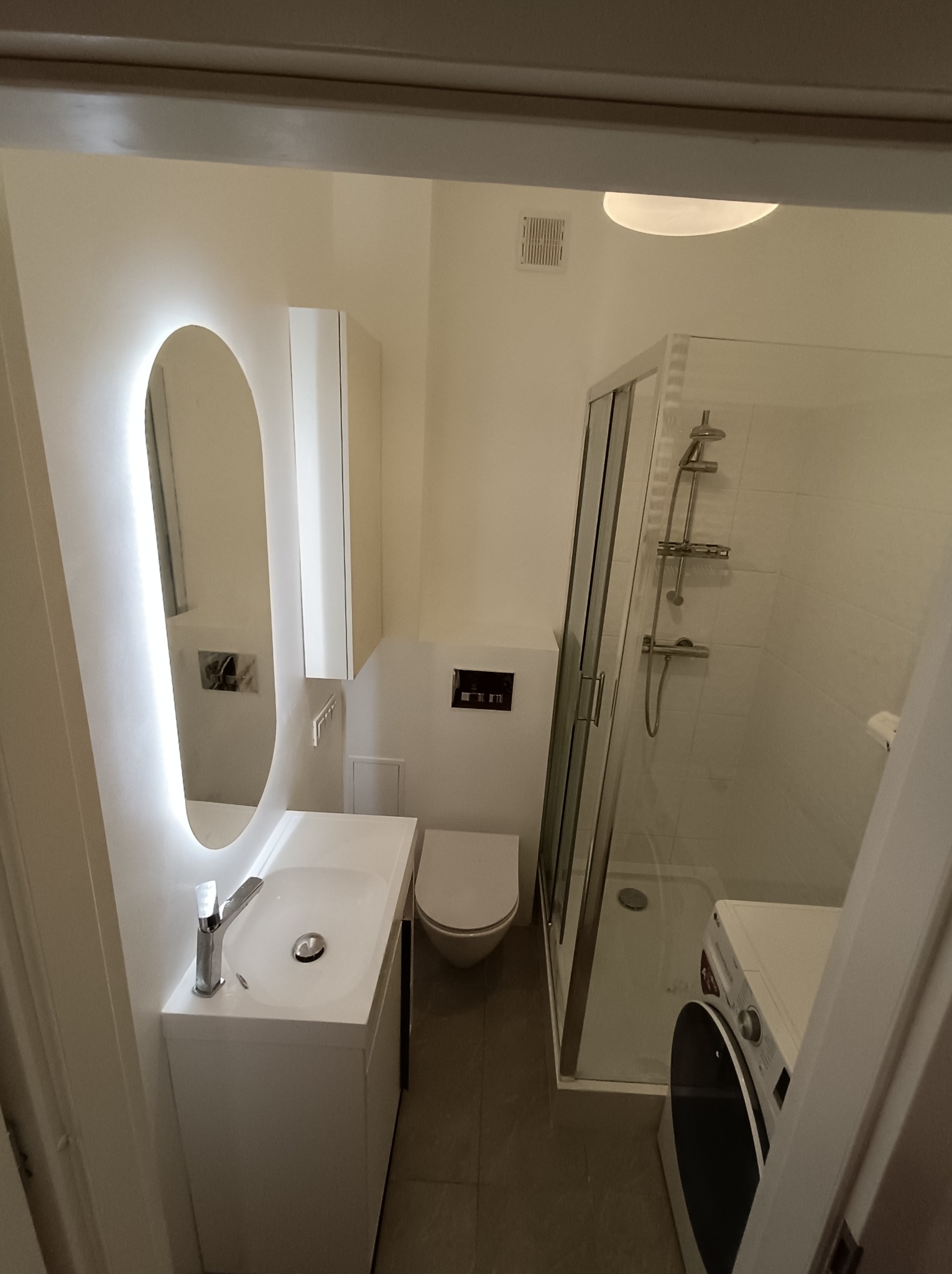 Jasna łazienka z owalnym lustrem LED, białą szafką z umywalką, kabiną prysznicową i pralką. Minimalistyczny design, neutralna kolorystyka.