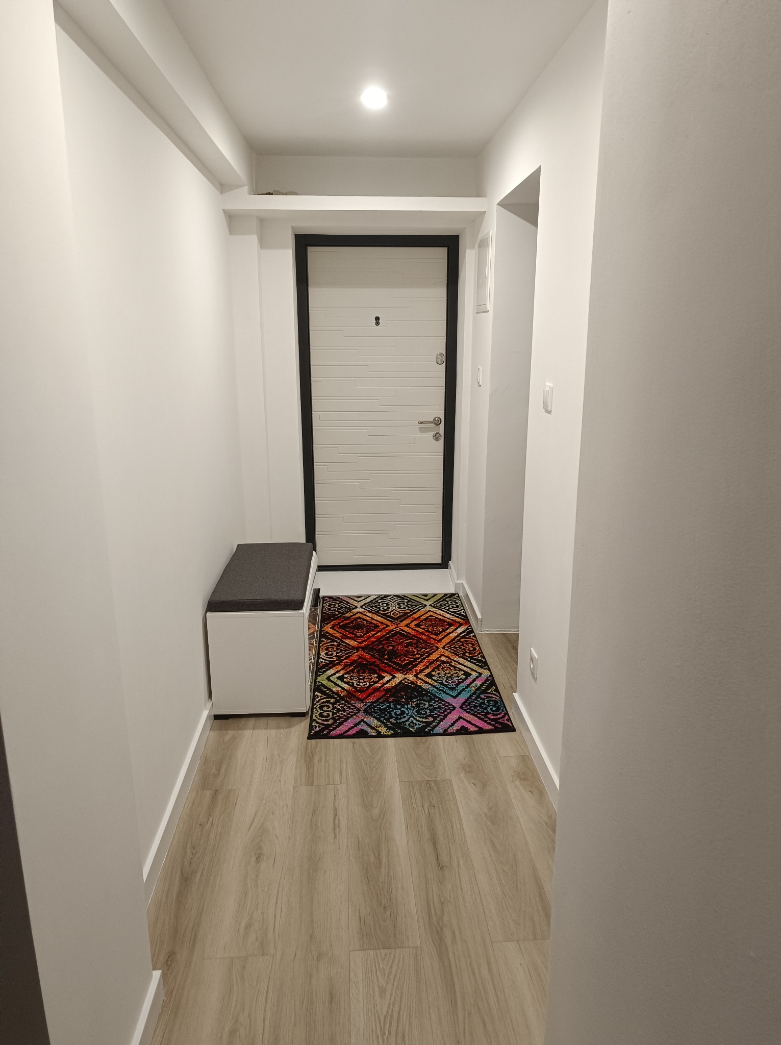 Jasny korytarz z panelami podłogowymi, białą ławką z szarym siedziskiem i kolorowym dywanikiem przed drzwiami wejściowymi z czarną ramą.