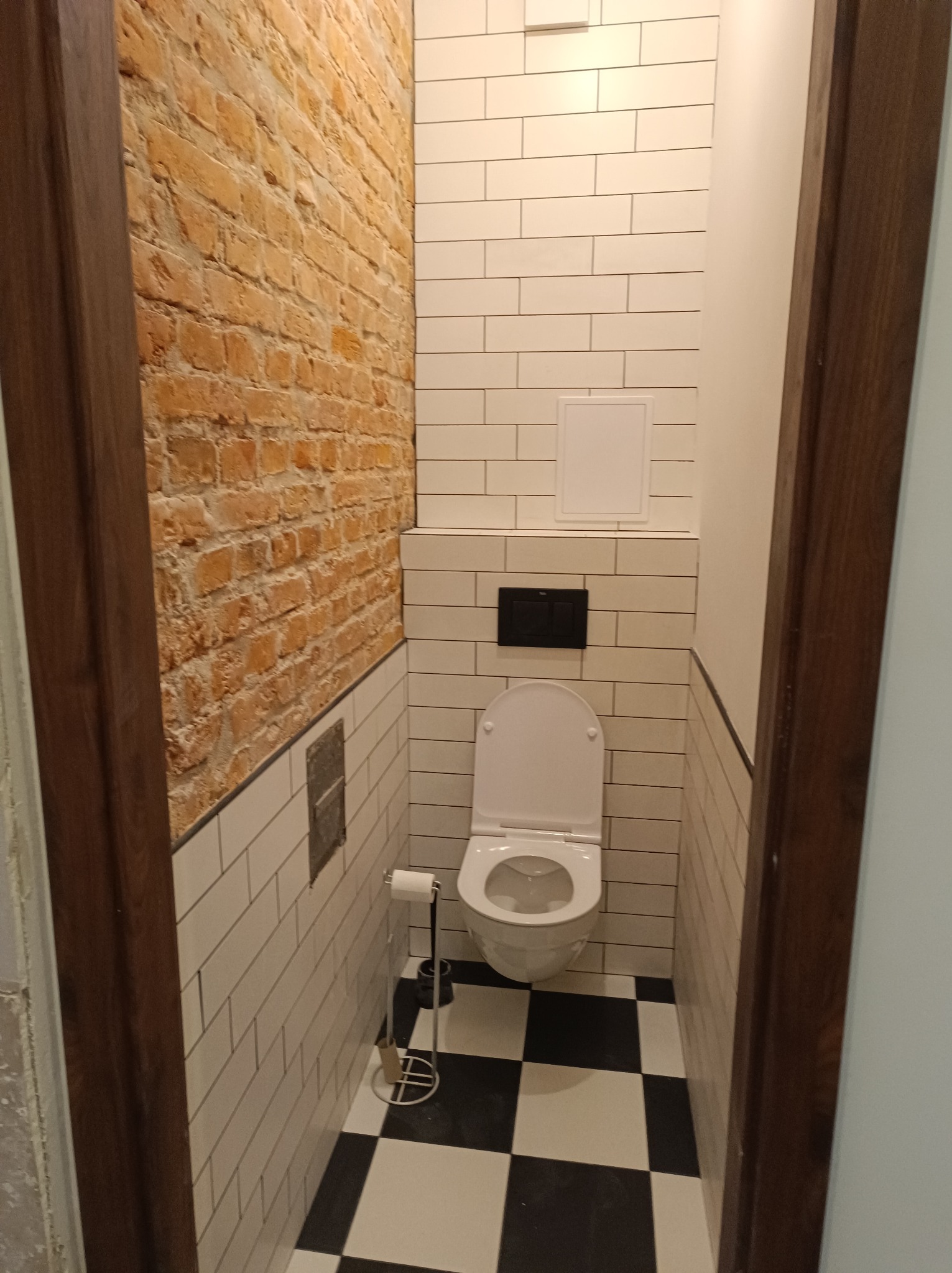 Wnętrze łazienki po remoncie: białe płytki, ściana z cegły, toaleta i czarno-biała podłoga w szachownicę. Widok zza futryny drzwi.