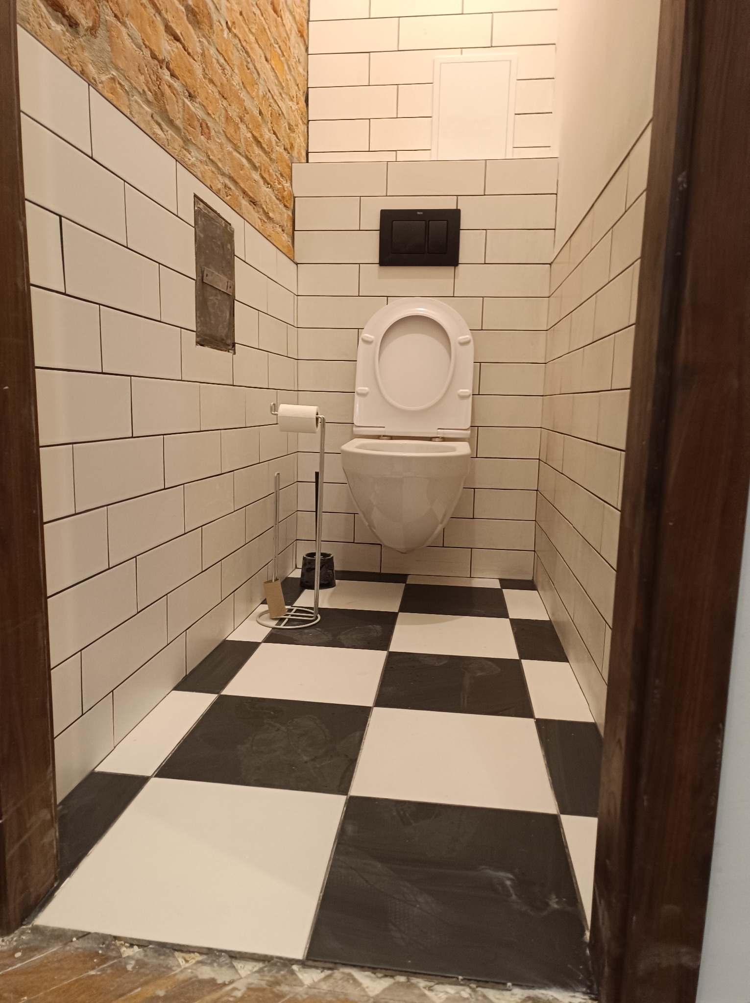 Ukończona łazienka z białą ceramiką, czarno-białą podłogą w szachownicę i fragmentem ściany z cegły. Widoczne detale wykończenia i akcesoria łazienkowe.