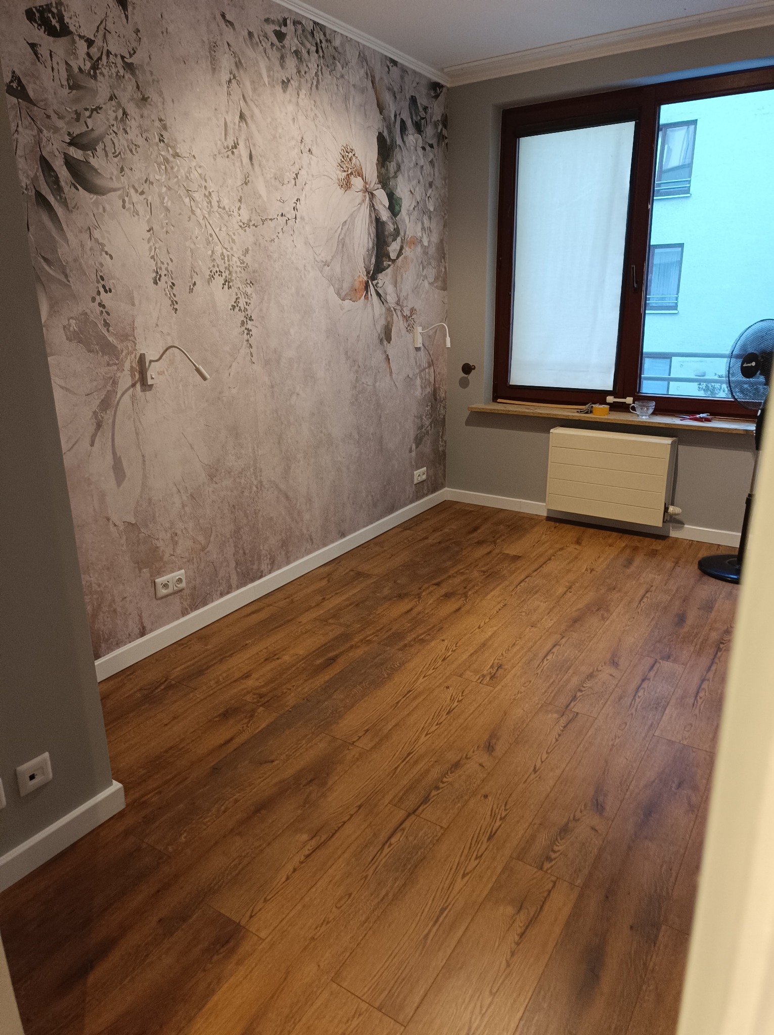 Pokój po remoncie: drewniana podłoga, ściana z fototapetą w kwiaty, okno z roletą. Widoczne gniazdka, listwy przypodłogowe i grzejnik. Minimalistyczny design.