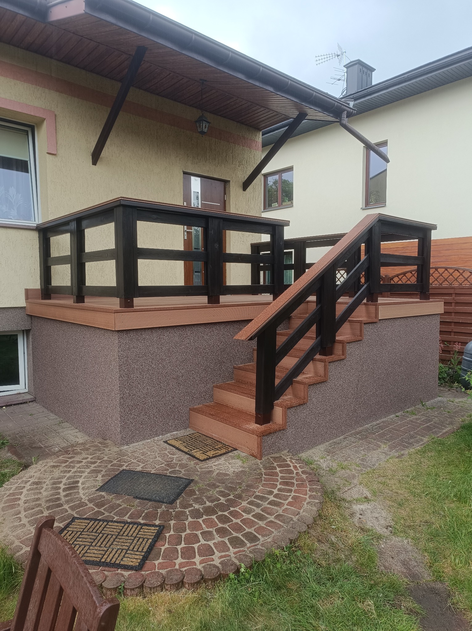 Zadaszony taras z drewnianą balustradą i schodami, wykończony brązowym kamieniem elewacyjnym. Widoczne wejście do budynku i fragment ogrodu z kostką brukową.