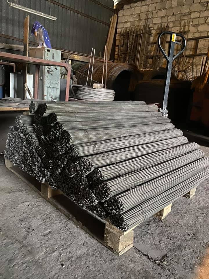 Paleta z wiązkami prętów żebrowanych w magazynie. Widoczne narzędzia i materiały budowlane w tle. Pręty ułożone równolegle, tworząc regularny wzór.