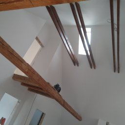 Dziadkowiec usługi ogólnobudowlane - Białe ściany poddasza z drewnianymi belkami stropowymi, widoczne okno i fragment drabiny malarskiej. Świeżo pomalowane wnętrze z zachowanymi elementami drewnianymi.