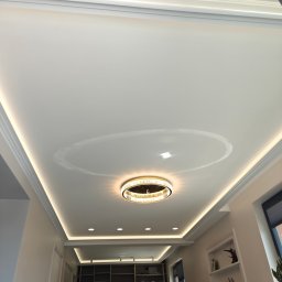 Dziadkowiec usługi ogólnobudowlane - Nowoczesny sufit z oświetleniem LED i dekoracyjnymi listwami. Okrągła lampa w centrum. Widok na wnętrze z półkami i oświetleniem.