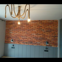 Dziadkowiec usługi ogólnobudowlane - Ściana z cegły klinkierowej z szarą boazerią i lampami ściennymi. Nad nią oryginalna lampa sufitowa z grubego sznura. Wnętrze po remoncie.