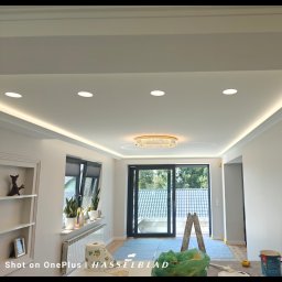 Dziadkowiec usługi ogólnobudowlane - Jasne wnętrze w trakcie prac wykończeniowych: zabezpieczona podłoga, drabina oparta o okno balkonowe, widoczne puszki z farbą. Oświetlenie sufitowe LED.