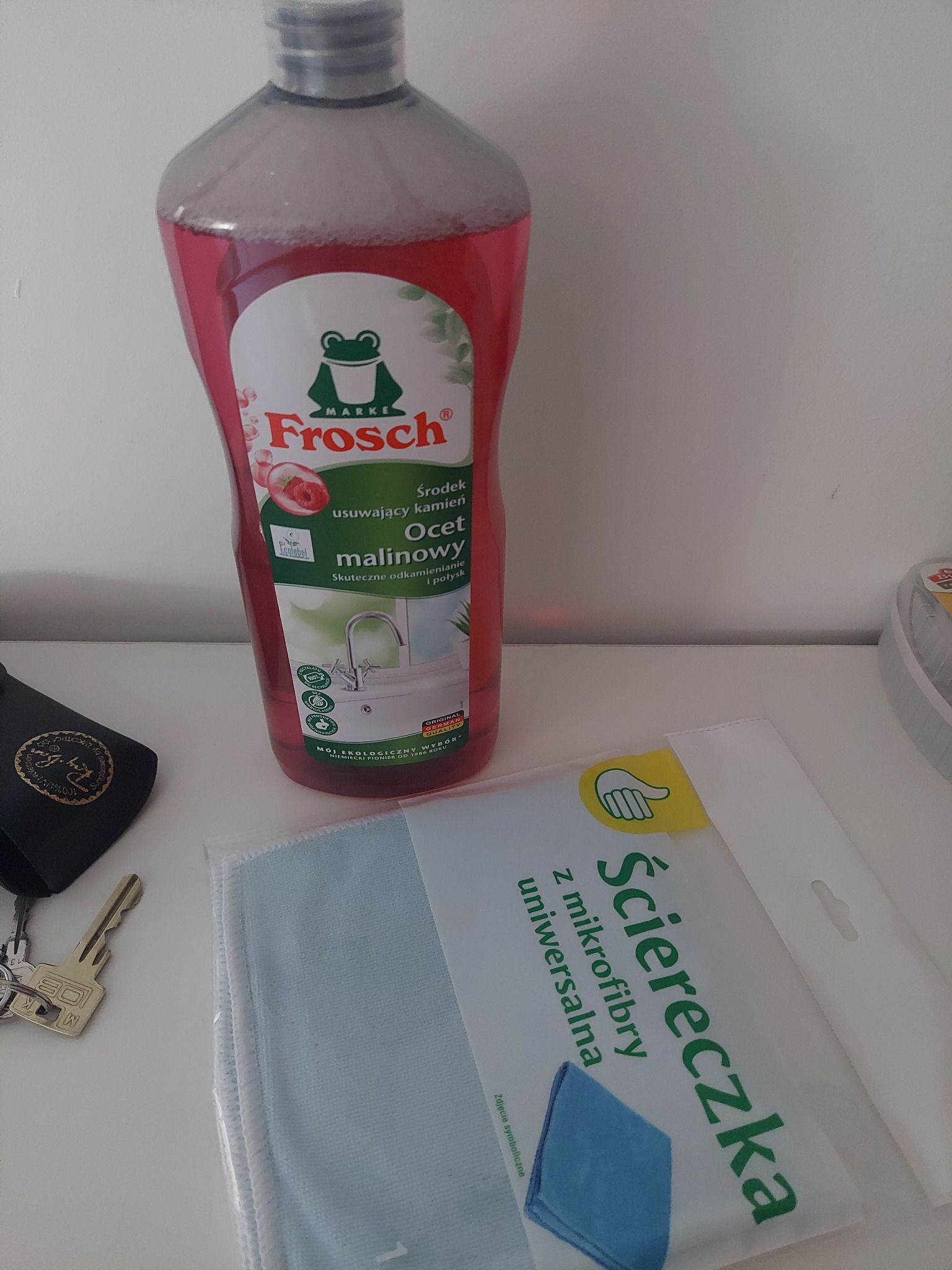 Butelka Frosch ocet malinowy, ściereczka z mikrofibry, klucze i etui okularów na białym blacie. Środek czyszczący i akcesoria do sprzątania.