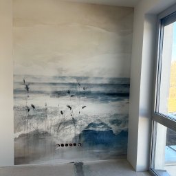 Fachowiec Group SP z o o - Ściana z fototapetą o motywie pejzażu morskiego w odcieniach bieli i błękitu, widoczne gniazdka elektryczne i surowa podłoga, okno z widokiem na zewnątrz.