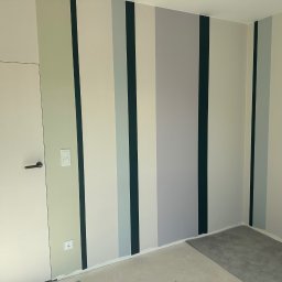 Fachowiec Group SP z o o - Ściana w pokoju pomalowana w pionowe pasy w odcieniach szarości, zieleni i bieli. Widoczna biała ściana z drzwiami i czarną klamką. Podłoga w trakcie prac wykończeniowych.