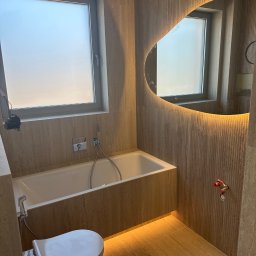 Fachowiec Group SP z o o - Nowoczesna łazienka z beżowymi płytkami, podświetlanym lustrem o nieregularnym kształcie i minimalistyczną armaturą. Widoczna wanna i toaleta.