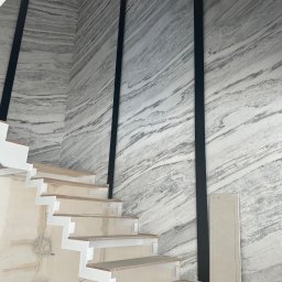 Fachowiec Group SP z o o - Montaż schodów z białymi wspornikami i drewnianymi stopniami na tle ściany z efektem marmuru, z czarnymi pionowymi elementami dekoracyjnymi. Prace wykończeniowe w toku.