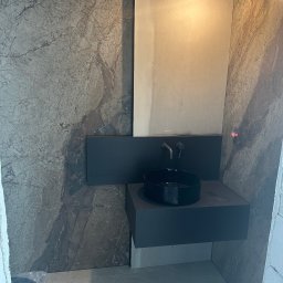 Fachowiec Group SP z o o - Nowoczesna łazienka z umywalką nablatową i marmurową okładziną ścienną. Minimalistyczny design, czarna armatura, surowe wykończenie. Widoczna żarówka na kablu.