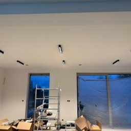 Fachowiec Group SP z o o - Białe wnętrze w trakcie wykańczania: zamontowane czarne lampy sufitowe, okna zabezpieczone folią, drabina i kartony na podłodze. Widoczne przewody elektryczne.