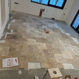 Fachowiec Group SP z o o - Układanie beżowych płytek podłogowych w jasnym wnętrzu. Widoczne narzędzia, plan pomieszczenia i okno z widokiem na zimowy krajobraz. Częściowo ułożona podłoga.