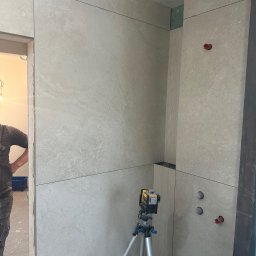 Fachowiec Group SP z o o - Wykończona ściana w łazience z beżowych płytek, widoczny laser krzyżowy na statywie, fragment sylwetki osoby. Minimalistyczny design, precyzyjne ułożenie płytek.