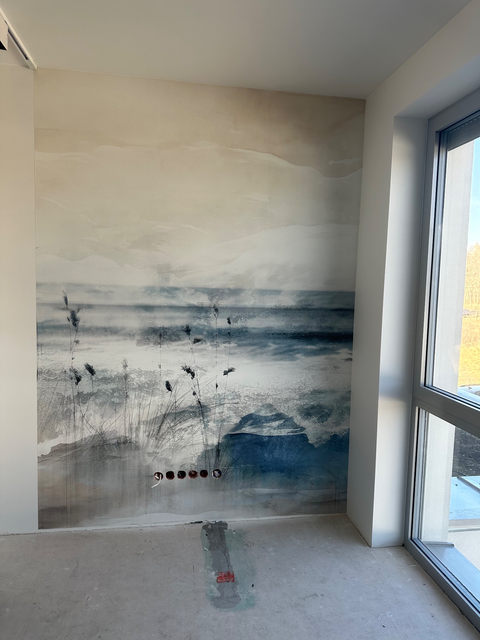Ściana z fototapetą o motywie pejzażu morskiego w odcieniach bieli i błękitu, widoczne gniazdka elektryczne i surowa podłoga, okno z widokiem na zewnątrz.