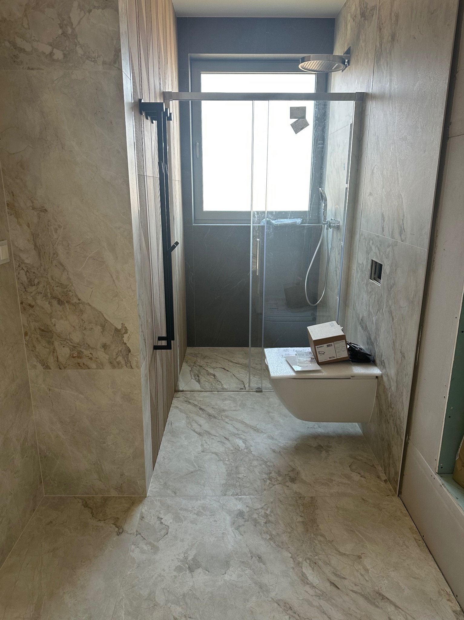 Wykończona łazienka z kabiną prysznicową typu walk-in, wiszącą toaletą i marmurowymi płytkami. Widoczne elementy montażowe i pudełko po produkcie.