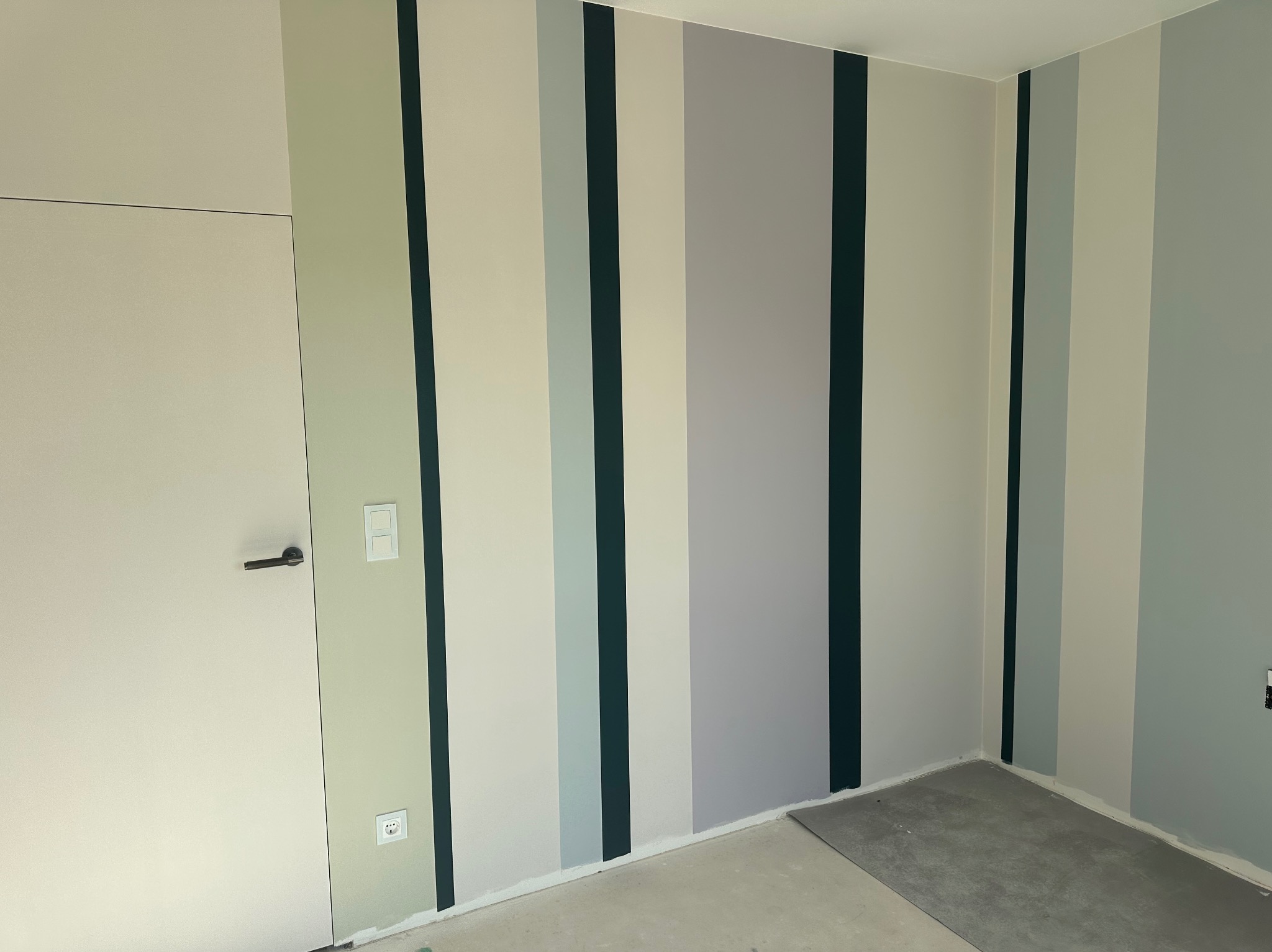 Ściana w pokoju pomalowana w pionowe pasy w odcieniach szarości, zieleni i bieli. Widoczna biała ściana z drzwiami i czarną klamką. Podłoga w trakcie prac wykończeniowych.