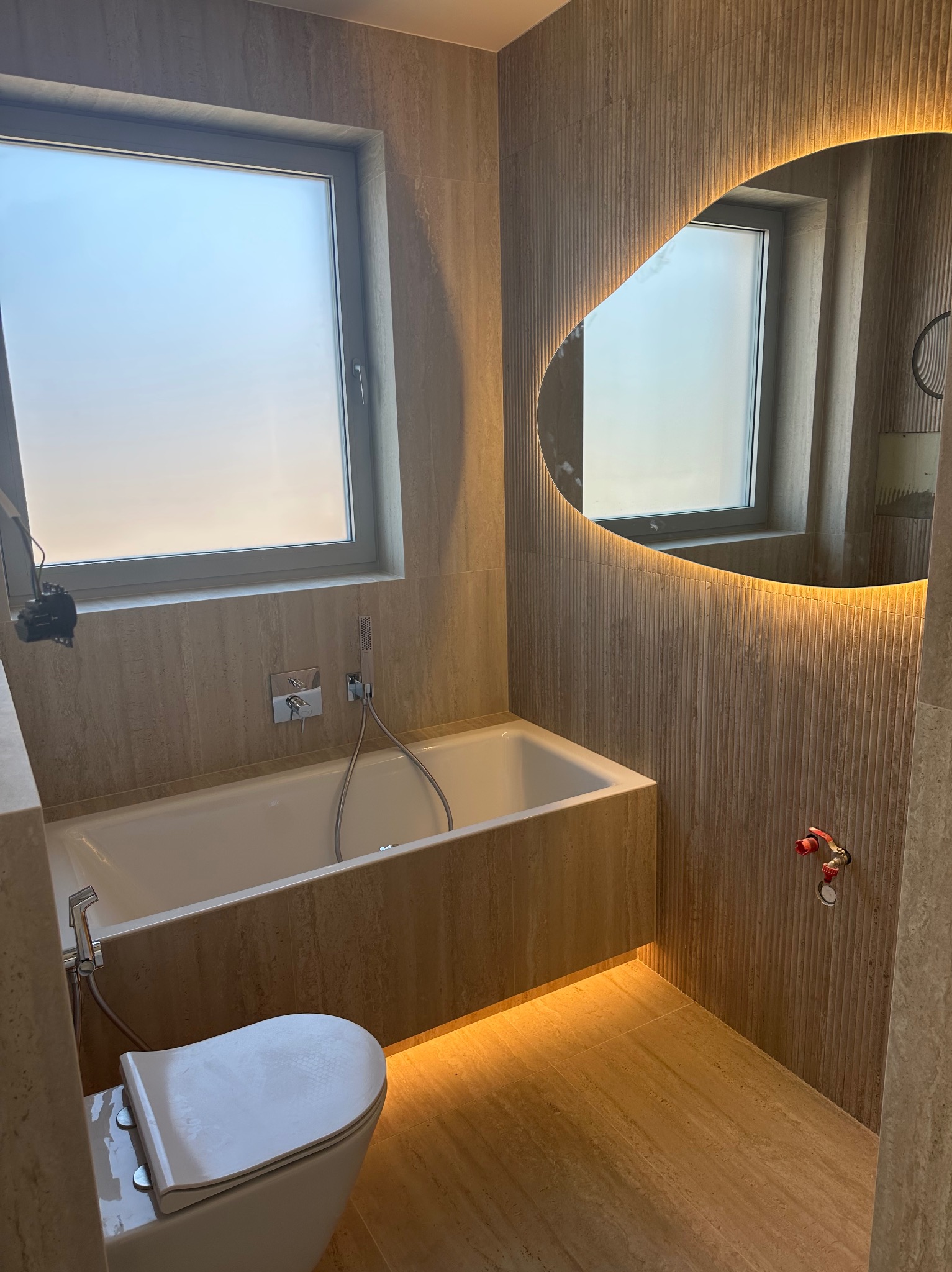 Nowoczesna łazienka z beżowymi płytkami, podświetlanym lustrem o nieregularnym kształcie i minimalistyczną armaturą. Widoczna wanna i toaleta.