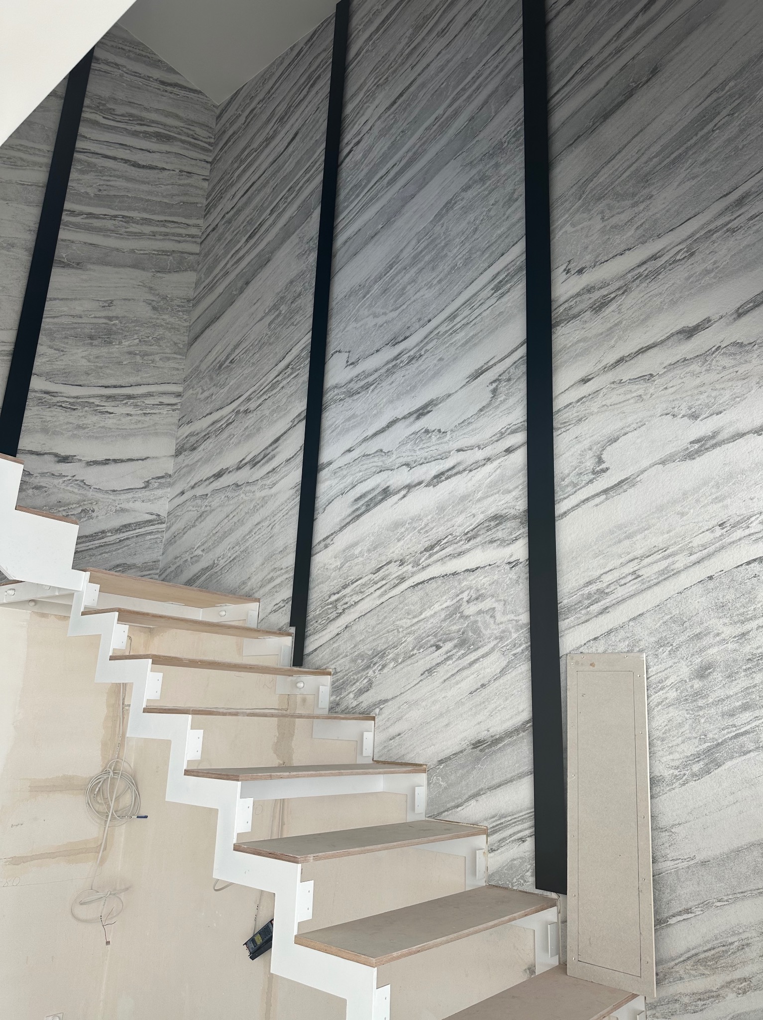 Montaż schodów z białymi wspornikami i drewnianymi stopniami na tle ściany z efektem marmuru, z czarnymi pionowymi elementami dekoracyjnymi. Prace wykończeniowe w toku.