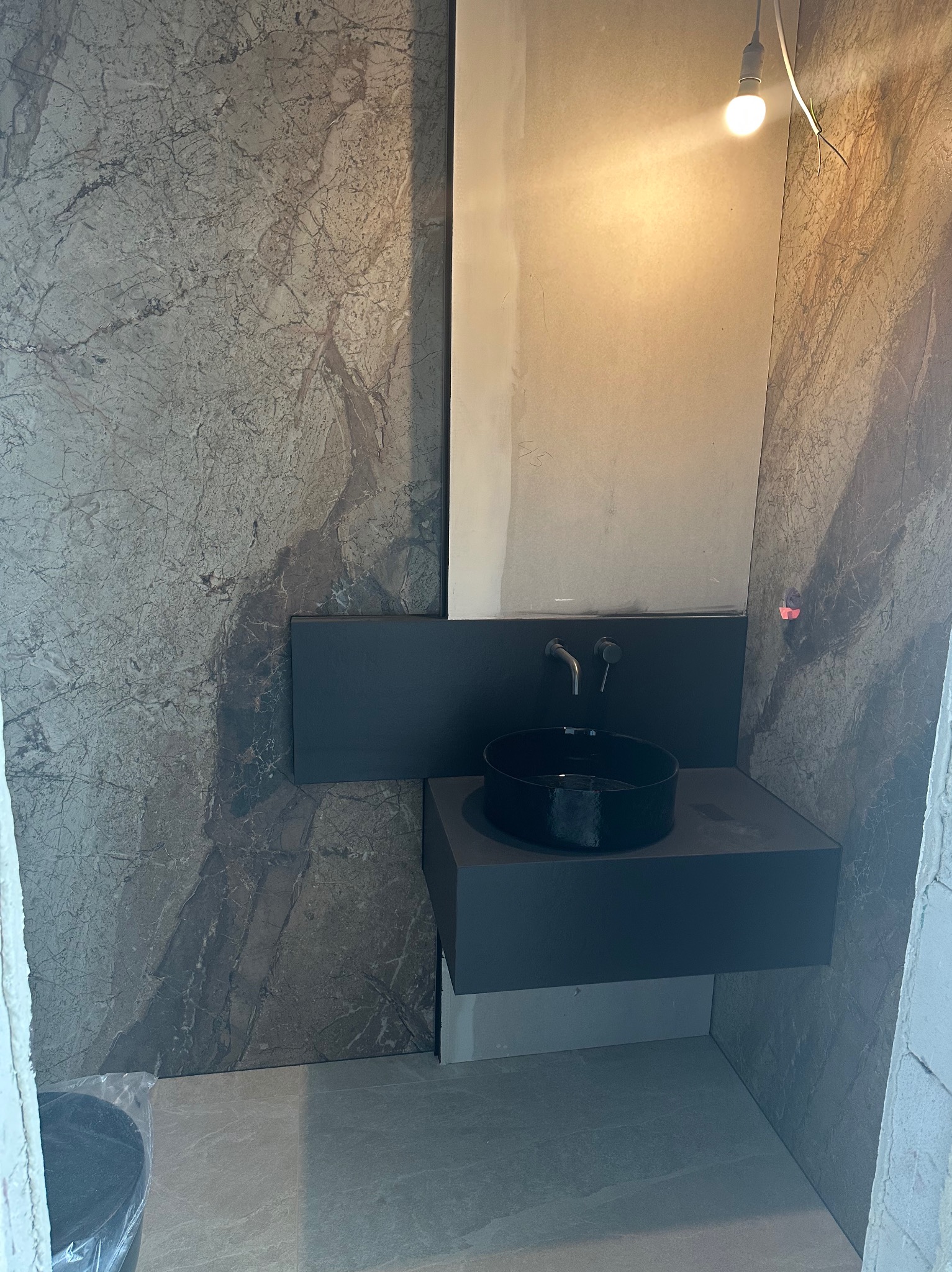 Nowoczesna łazienka z umywalką nablatową i marmurową okładziną ścienną. Minimalistyczny design, czarna armatura, surowe wykończenie. Widoczna żarówka na kablu.