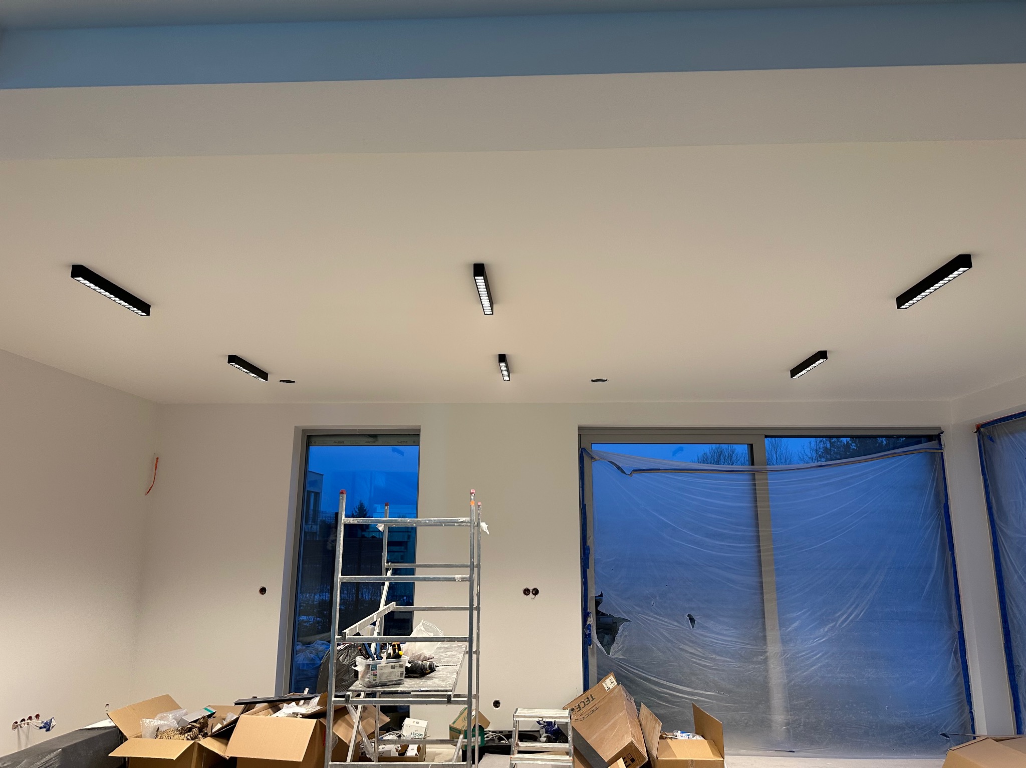 Białe wnętrze w trakcie wykańczania: zamontowane czarne lampy sufitowe, okna zabezpieczone folią, drabina i kartony na podłodze. Widoczne przewody elektryczne.