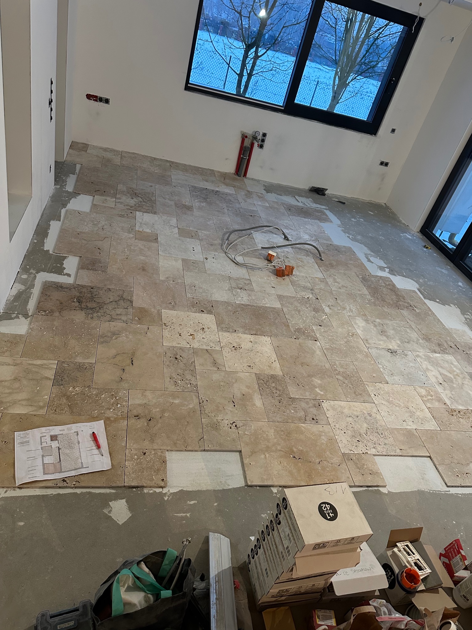 Układanie beżowych płytek podłogowych w jasnym wnętrzu. Widoczne narzędzia, plan pomieszczenia i okno z widokiem na zimowy krajobraz. Częściowo ułożona podłoga.