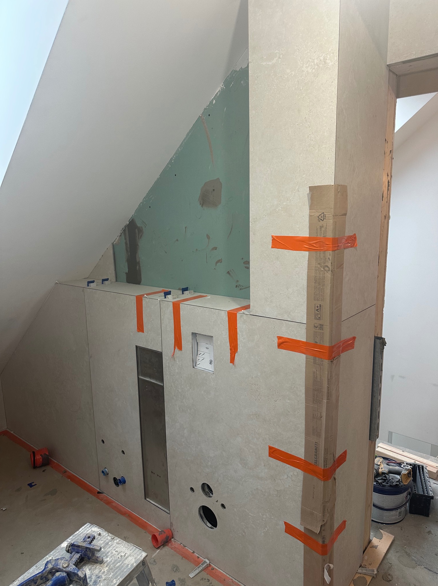 Montaż płytek w łazience na poddaszu, widoczne narzędzia, taśmy zabezpieczające i fragment ściany z płyt karton-gips. Etap wykończeniowy z widocznymi instalacjami.