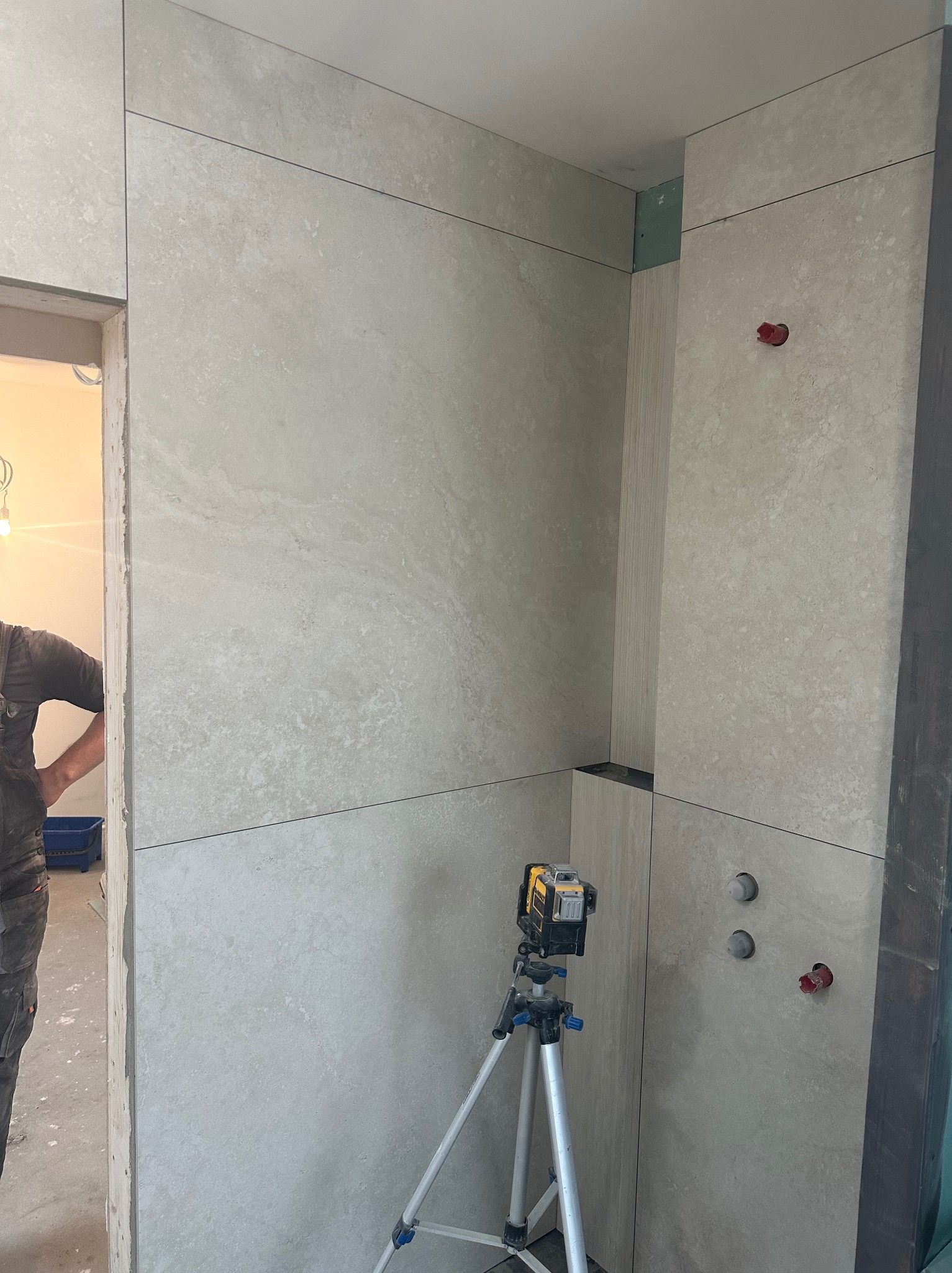 Wykończona ściana w łazience z beżowych płytek, widoczny laser krzyżowy na statywie, fragment sylwetki osoby. Minimalistyczny design, precyzyjne ułożenie płytek.