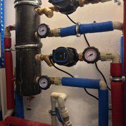 Bohdan Unhul - Instalacja grzewcza z czarnym zbiornikiem, pompami obiegowymi i manometrami. Rury w izolacji czerwonej i niebieskiej. Widoczne zawory i złączki.