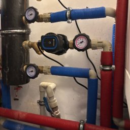 Bohdan Unhul - Fragment instalacji hydraulicznej z czarnym filtrem, manometrami i pompami obiegowymi, rury w izolacji niebieskiej i czerwonej, widoczne złącza i zawory.