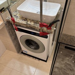 Bohdan Unhul - Montaż umywalki nablatowej na pralce w łazience, zabezpieczonej ściskami stolarskimi. Blat z granitu, nowoczesna armatura, płytki w odcieniach beżu i brązu.