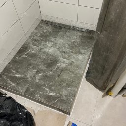 Bohdan Unhul - Wykończony brodzik z szarych płytek imitujących marmur, z odpływem liniowym. Białe płytki na ścianach, widoczny fragment podłogi z jasnych płytek. Częściowo zakryty folią.