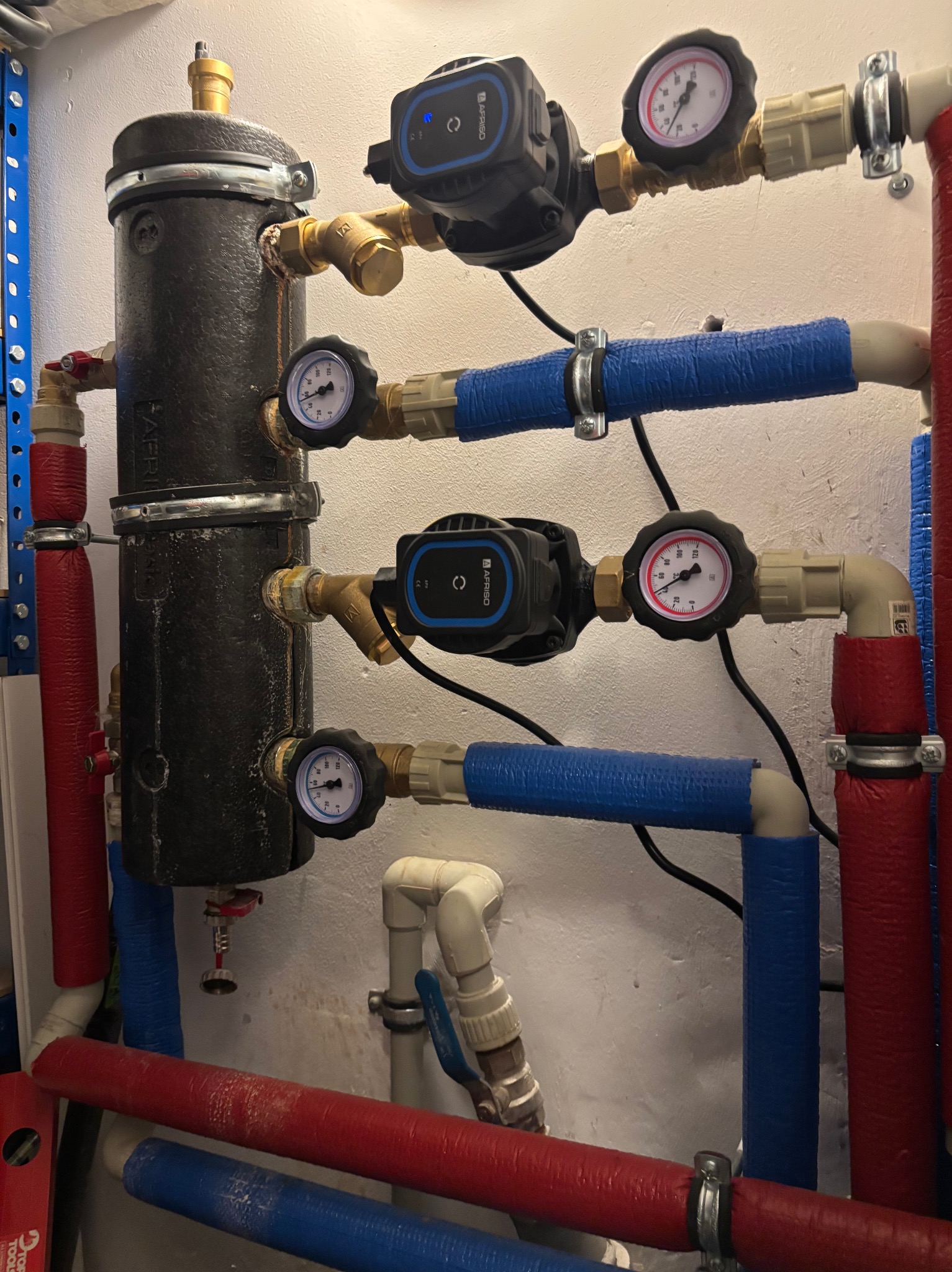 Instalacja grzewcza z czarnym zbiornikiem, pompami obiegowymi i manometrami. Rury w izolacji czerwonej i niebieskiej. Widoczne zawory i złączki.