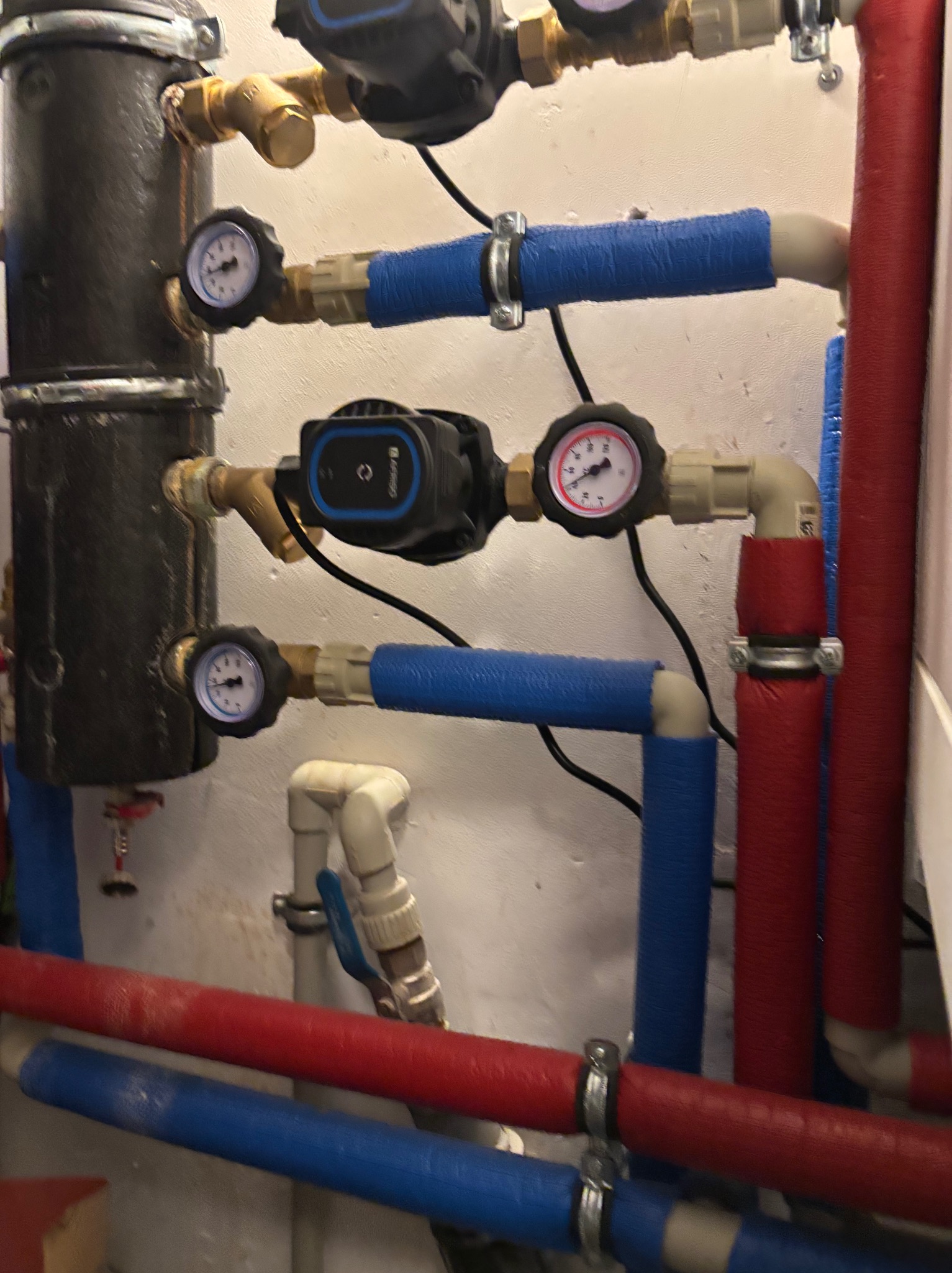 Fragment instalacji hydraulicznej z czarnym filtrem, manometrami i pompami obiegowymi, rury w izolacji niebieskiej i czerwonej, widoczne złącza i zawory.