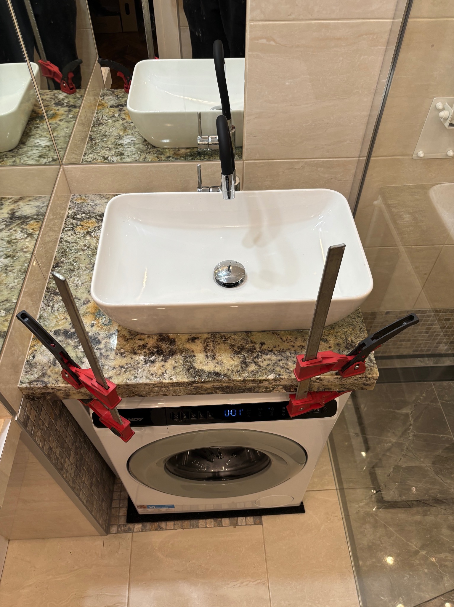 Montaż umywalki nablatowej na pralce w łazience, zabezpieczonej ściskami stolarskimi. Blat z kamienia imitującego granit. Widok z góry na armaturę i ceramikę.