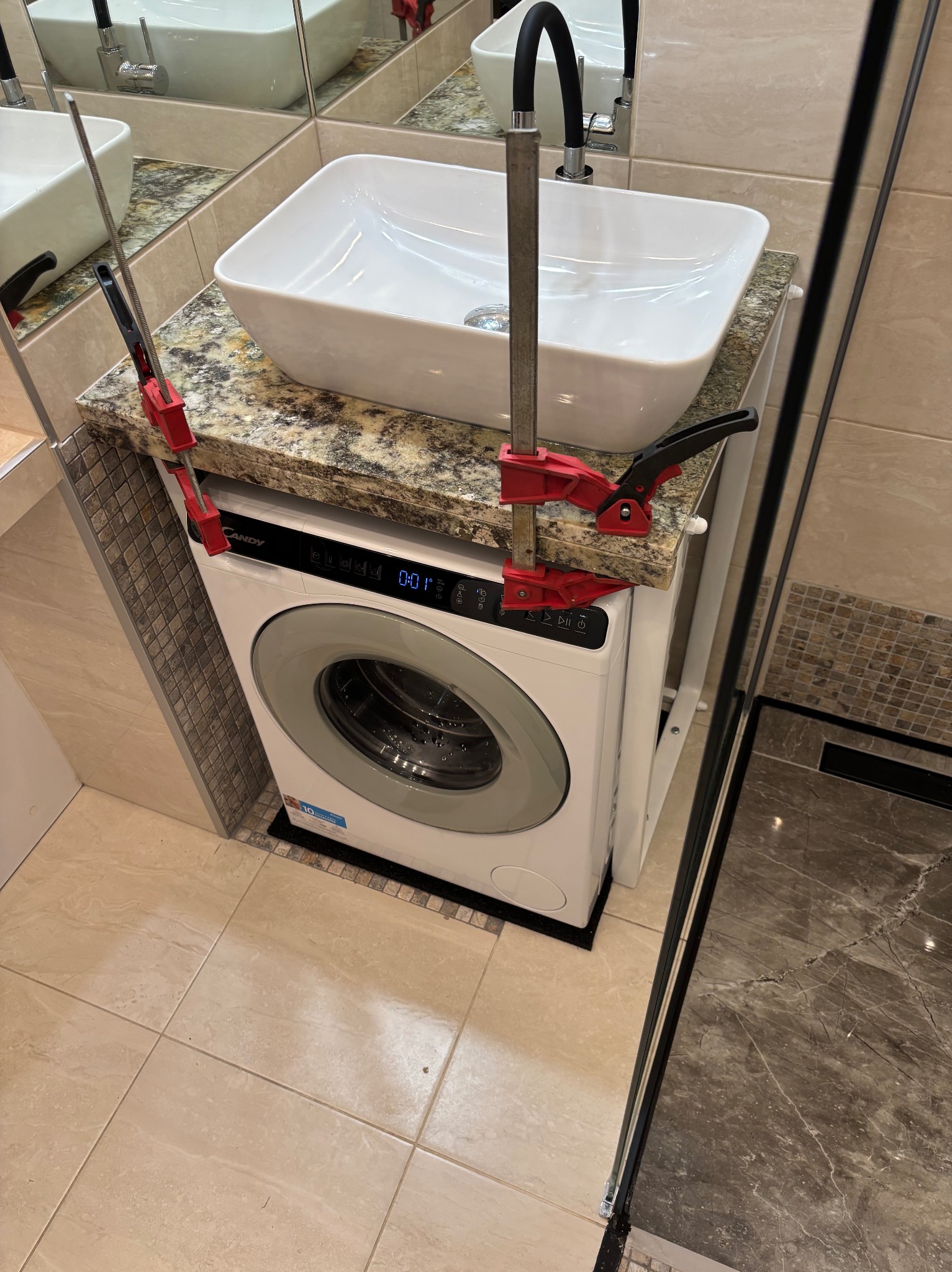 Montaż umywalki nablatowej na pralce w łazience, zabezpieczonej ściskami stolarskimi. Blat z granitu, nowoczesna armatura, płytki w odcieniach beżu i brązu.