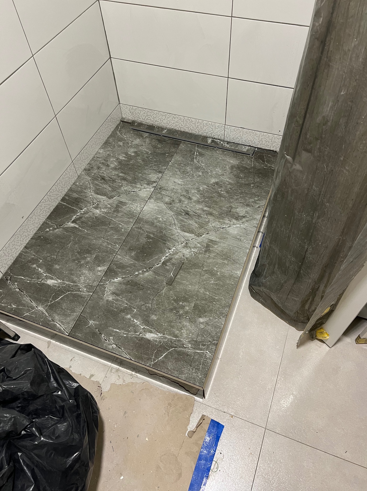 Wykończony brodzik z szarych płytek imitujących marmur, z odpływem liniowym. Białe płytki na ścianach, widoczny fragment podłogi z jasnych płytek. Częściowo zakryty folią.