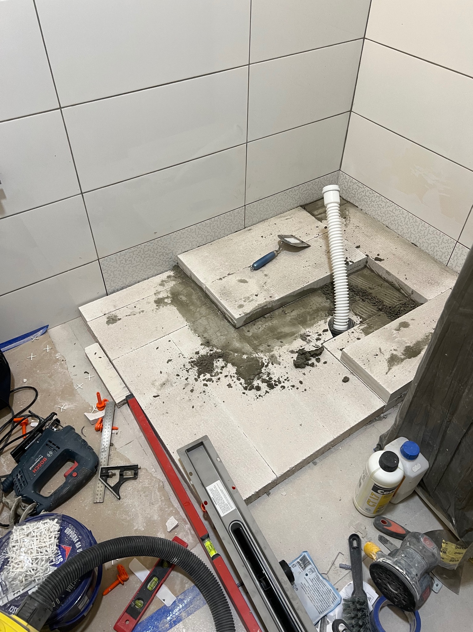 Montaż odpływu liniowego w kabinie prysznicowej. Widoczne narzędzia, białe płytki na ścianach i częściowo ułożone płyty podłogowe z otworem na rurę odpływową.