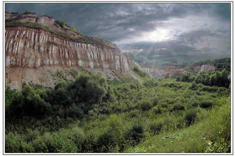 Rozległy krajobraz Ukrainy z erodowanymi klifami o warstwowej strukturze, porośniętymi zielenią w dolinie, pod pochmurnym niebem z promieniami słońca.