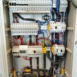 MICHAŁ Z NAPIĘCIEM MICHAŁ PAWLAK - Rozdzielnica elektryczna w obudowie, widoczne zabezpieczenia, przewody, listwy zaciskowe i narzędzia monterskie na dnie szafy. Instalacja wykonana schludnie, przewody uporządkowane.