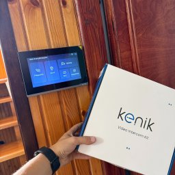 MICHAŁ Z NAPIĘCIEM Michał Pawlak - Zamontowany panel wideodomofonu Kenik na drewnianej ścianie, obok pudełko z zestawem. Widoczny fragment ręki z zegarkiem. Ustawienia: połączenie, drzwi, kamery.